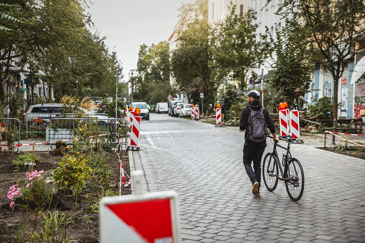 Eine Person läuft mit einem Fahrrad auf einer teilweise gesperrten Straße, umgeben von Bauabsperrungen und Grünanlagen.